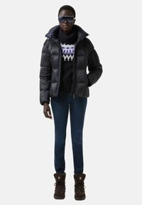 Vatelínová černá bunda, pletený černý svetr s bílým a fialovým vzorem, modré džíny a hnědé boty s kožešinovým lemováním a detailem loga.