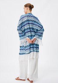 Blauwe en witte sjaal met zigzagpatroon en franjes. Draag het over een witte broek en sandalen voor een ontspannen, gelaagde uitstraling.
