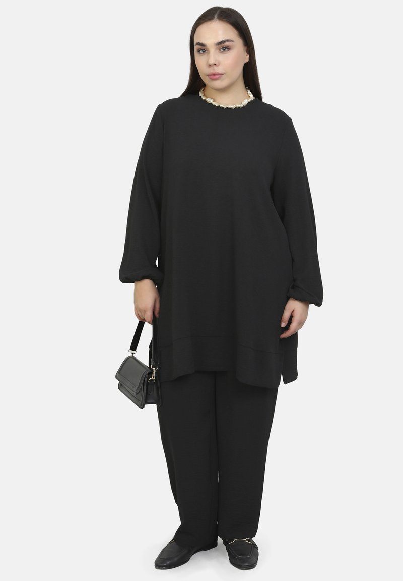 Sort oversized sweater med lange ærmer, parret med matchende vide bukser. Holder en lille sort håndtaske og har flade sko på. Neutral baggrund.