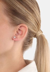 Kvinna med blont hår uppsatt som bär en silvrig blommformad örhänge med klara ädelstenar i vänster öra.