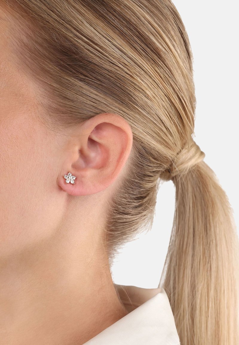 Kvinna med blont hår uppsatt som bär en silvrig blommformad örhänge med klara ädelstenar i vänster öra.