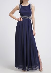 Frau mit einem ärmellosen, marineblauen Maxikleid mit verzierten Trägern und Taillendetails, kombiniert mit beigen, offenen Sandalen.