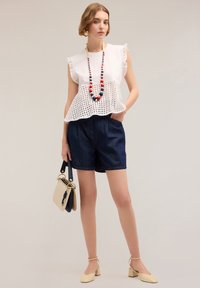 Blusa blanca de ojales con mangas rizadas, pantalones cortos de mezclilla oscura y un bolso beige. Los accesorios incluyen un collar de cuentas coloridas y zapatos de tacón cuadrado.