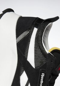 Sorte og hvite sneakers med mesh-overdel og semsket detaljer, teksturerte materialer, avrundet form og gule innervaler.