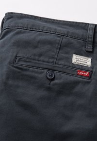Mørkegrå bukser med denimstruktur, med en baglomme, en sort knap og en hvid stofmærke med røde accenter.