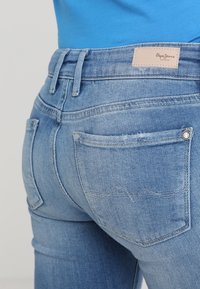 Vaqueros de denim azul claro con una cintura alta, dos bolsillos traseros con un diseño de costura en forma de ola y una etiqueta de cuero con marca en la cinturilla.