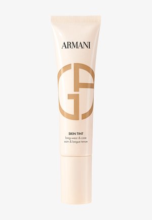 Armani Skin Tint in un tubetto beige e lucido con un logo dorato. Presenta un applicatore a pressione e un testo che evidenzia i benefici del prodotto sottostante.