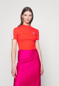 Top corto rosso a coste con collo alto e maniche corte, con dettagli bianchi e un piccolo logo. Indossato con una gonna rosa lucida.