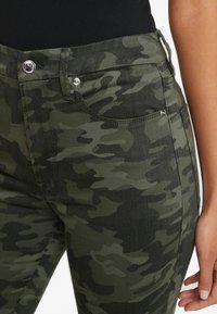 Jeans de talle alto con patrón de camuflaje en tonos verdes. Presentan un cierre con cremallera, bolsillos frontales y detalles en metal plateado. Tejido texturizado.