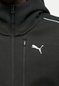 Κοντινή λήψη ενός μαύρου ζακέτου με φερμουάρ, με μια μικρή τσέπη με φερμουάρ στο στήθος και λευκό λογότυπο Puma στη δεξιά πλευρά.