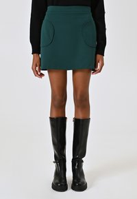 Mini gonna verde con vita alta, due tasche tonde, realizzata in un tessuto morbido. Abbinata a stivali neri al ginocchio con cerniere.