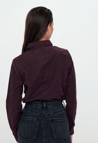 Camicia bordeaux a maniche lunghe con colletto, realizzata in un tessuto testurizzato. Indossata con una gonna di jeans nera a vita alta, vista da dietro.