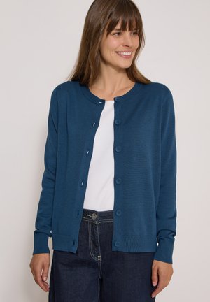Femme souriante portant un cardigan bleu boutonné par-dessus une chemise blanche et un jean foncé, debout devant un fond clair uni.