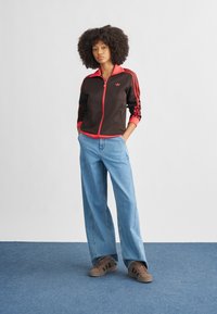 Brown zip-up jacket with red accents and three stripes on sleeves, paired with light blue wide-leg jeans and brown sneakers.