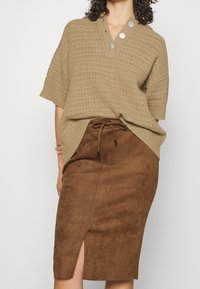 Pull en tricot beige à manches courtes avec trois boutons décoratifs au col, associé à une jupe midi en daim marron avec une fente sur le côté.