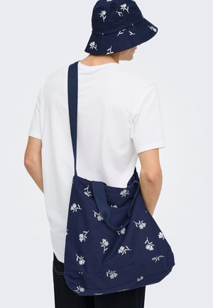 Personne portant un t-shirt blanc, un chapeau bob bleu marine et un sac à bandoulière bleu marine assorti avec des motifs floraux blancs, vue de dos.