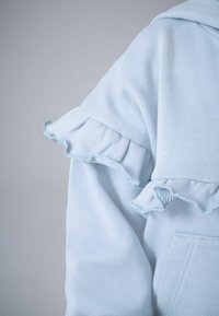 Hanorac albastru deschis, cu detalii plisate pe umeri, țesătură moale și texturată și buzunar frontal.