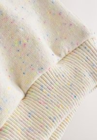Primer plano de una tela crema clara con pequeñas motas multicolores y detalle de puño acanalado.