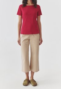 Nainen, jolla on päällään lyhythihainen punainen paita, beige leveälahkeiset pussihousut ja oliivinvihreät liukusandalit, seisoo yksinkertaista taustaa vasten.