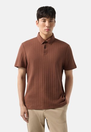 Ung mand iført en brun ribbet kortærmet polotrøje og beige bukser, stående foran en ensfarvet lys baggrund.