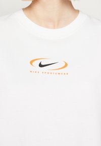 Vit T-shirt med kort ärm och rund halsringning, prydd med en svart Nike-logotyp omgiven av en orange swoosh och texten "NIKE SPORTSWEAR."