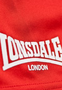 Crvena pamučna tkanina s bijelim tekstom koji glasi "LONSDALE" sa zakrivljenim dizajnom, a ispod u manjem fontu "LONDON".