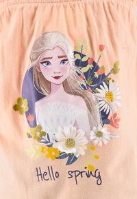 Tessuto pesca chiaro con un'immagine di un personaggio circondato da fiori. Il testo sottostante recita "Ciao primavera." Presenta accenti dorati e colori vivaci.