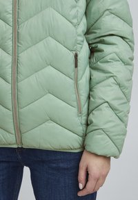 Veste matelassée vert clair avec un design en chevrons, dotée d'une fermeture éclair à l'avant et de poches latérales, fabriquée en matériau lisse et texturé.