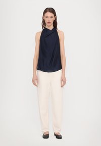 Navy silk halterneck-top laskeutuu pehmeästi, yhdistettynä kermaisiin leveälahkeisiin housuihin; mustat flatsit täydentävät asua. Yksinkertainen ja tyylikäs muotoilu.