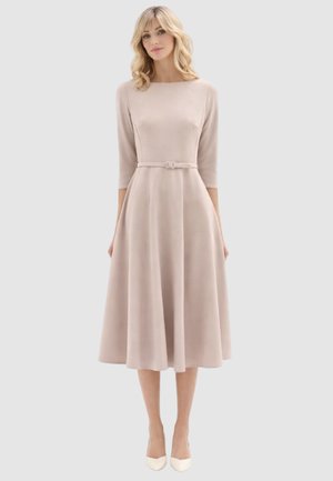 Donna che indossa un vestito midi beige a maniche lunghe con cintura aderente in vita e décolleté bianchi con punta a punta, in piedi davanti a uno sfondo semplice.