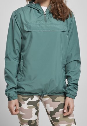 Personne portant une veste coupe-vent à capuche turquoise avec une demi-fermeture éclair et des leggings à motif camouflage, visible des épaules jusqu'au milieu des cuisses.