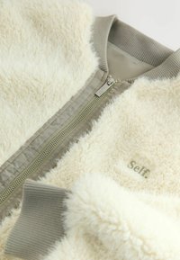 Kremowa kurtka bomberka o miękkiej, puszystej fakturze i oliwkowozielonych akcentach. Posiada pełny zamek błyskawiczny oraz ribbowe mankiety. Widoczne logo "Self.".