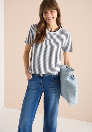 Kvinde iført en hvid og sortstribet kortærmet skjorte og blå jeans, der holder en lyseblå totesæk i venstre hånd.