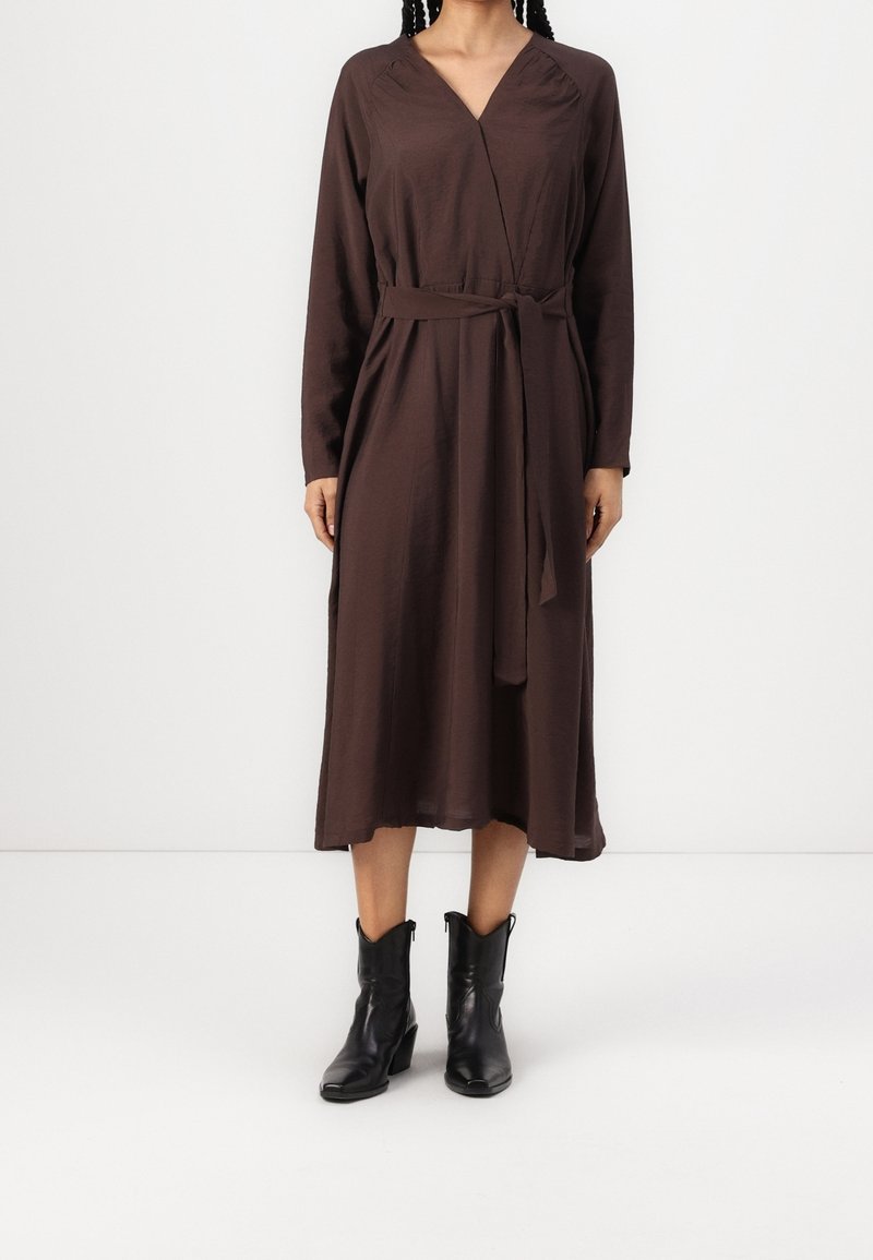 Femme portant une robe portefeuille marron foncé à manches longues avec une ceinture nouée et des bottes noires mi-mollet, debout devant un fond uni.