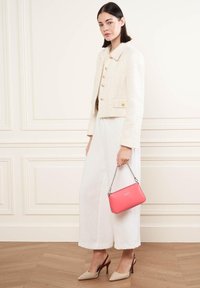 Giacca di crema texture con bottoni dorati, abbinata a pantaloni bianchi svolazzanti. Tiene una borsa rosa con tracolla a catena. Scarpe con tacco beige.
