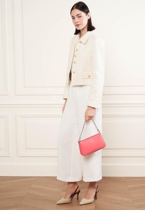Giacca di crema texture con bottoni dorati, abbinata a pantaloni bianchi svolazzanti. Tiene una borsa rosa con tracolla a catena. Scarpe con tacco beige.