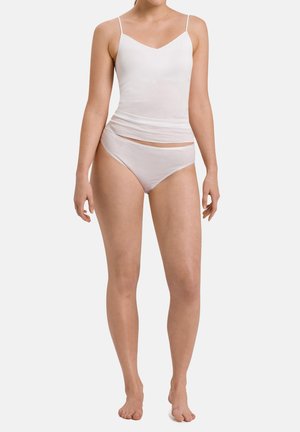 Frau steht barfuß und trägt ein weißes ärmelloses Top und passende Baumwollunterwäsche vor einem schlichten, hellen Hintergrund.