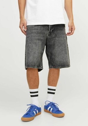 Persona que lleva puestos unos pantalones cortos de mezclilla negros, calcetines blancos con rayas negras, zapatillas azules con rayas blancas y una camisa blanca, de pie sobre un suelo blanco.