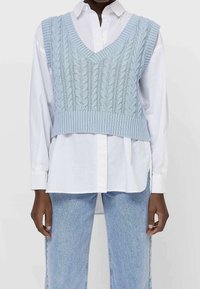 Osoba ubrana w jasnoniebieski sweter bez rękawów w splot kablowy, nosząca białą koszulę z guzikami oraz jasnoniebieskie dżinsy.