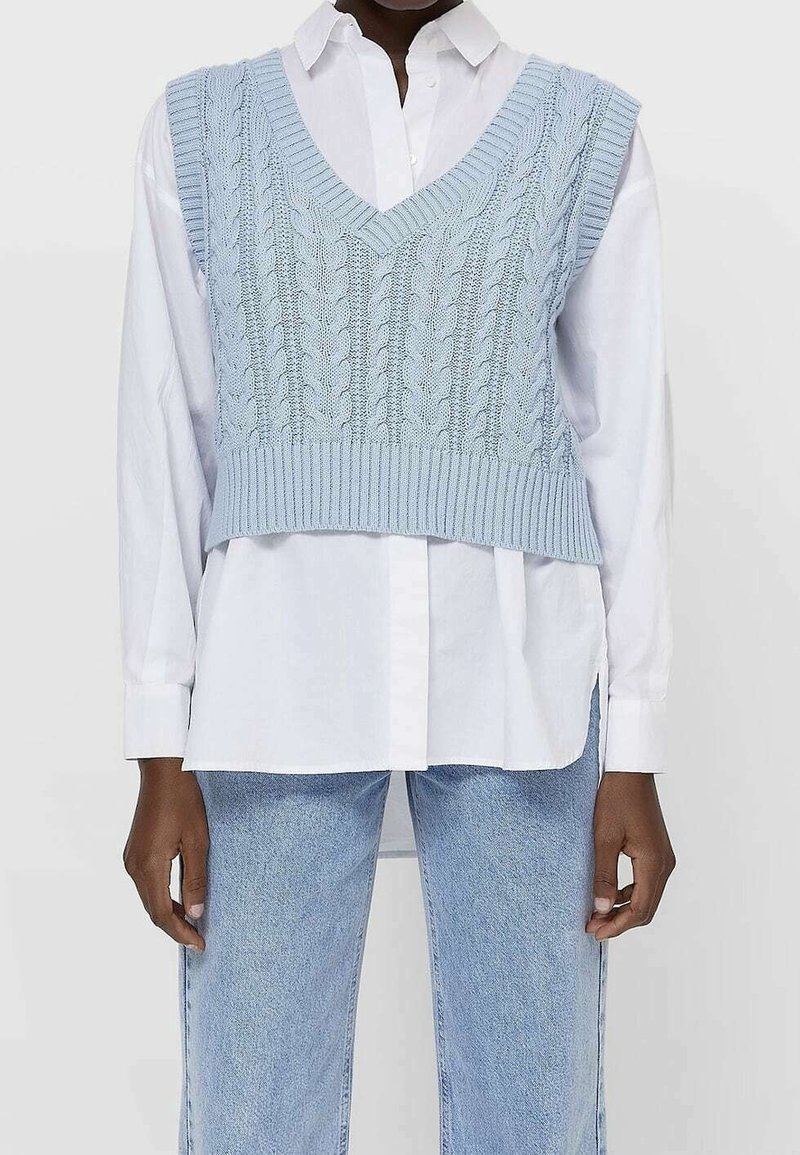 Osoba ubrana w jasnoniebieski sweter bez rękawów w splot kablowy, nosząca białą koszulę z guzikami oraz jasnoniebieskie dżinsy.