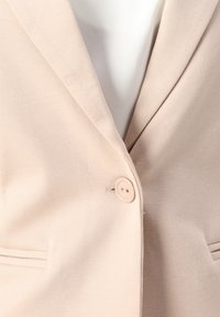 Blazer beige avec une fermeture à un seul bouton sur un vêtement intérieur blanc, montrant les revers et le bord supérieur des poches avant.