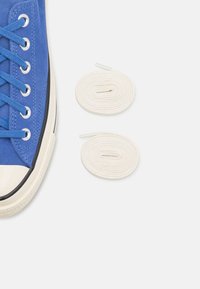 Blue suede sneaker with white rubber toe cap and black sole, featuring blue laces. Two white cotton shoelaces are positioned nearby.