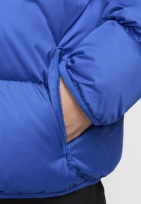 Mėlynas pūkinis švarkas su užtrauktuku kišenėje, pasižymintis lygia, blizgia tekstūra ir įstrižomis siūlėmis ant rankovės.