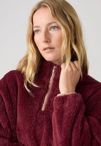 Bordo flis pulover z visokim ovratnikom, sprednjim zapenjanjem na pol zadrge in mehko, teksturirano tkanino. Ima oprijete manšete.