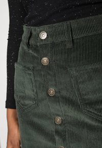 Jupe en velours côtelé vert avec texture côtelée verticale. Elle comporte des boutons en métal et des poches latérales. Un haut foncé contraste avec la couleur et la texture de la jupe.