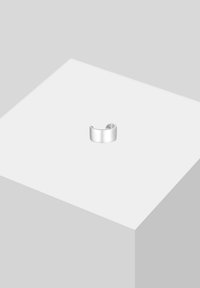 Bague manchette en argent lisse posée sur un cube blanc avec un fond gris clair, mettant en valeur son design minimaliste et sa forme ouverte.