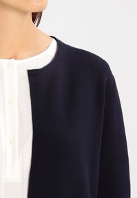 Marinefarbener Cardigan mit rundem Halsausschnitt, strukturiertem Stoff und offener Vorderseite, getragen über einem weißen Hemd mit Knopfschluss.