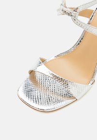 Sandale métallique argentée à bout ouvert avec motif texturé, bride à la cheville et semelle intérieure beige sur fond blanc.