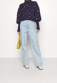 Pull en maille bleu marine avec pompons colorés, manches décontractées, associé à un jean large bleu clair et un sac bandoulière jaune. Chaussures blanches.