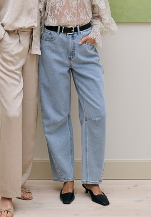 Zwei Personen stehen nebeneinander, eine trägt cremefarbene Hosen und beige Sandalen, die andere hellblaue Jeans, schwarzen Gürtel, Spitzenbluse und schwarze Absätze.